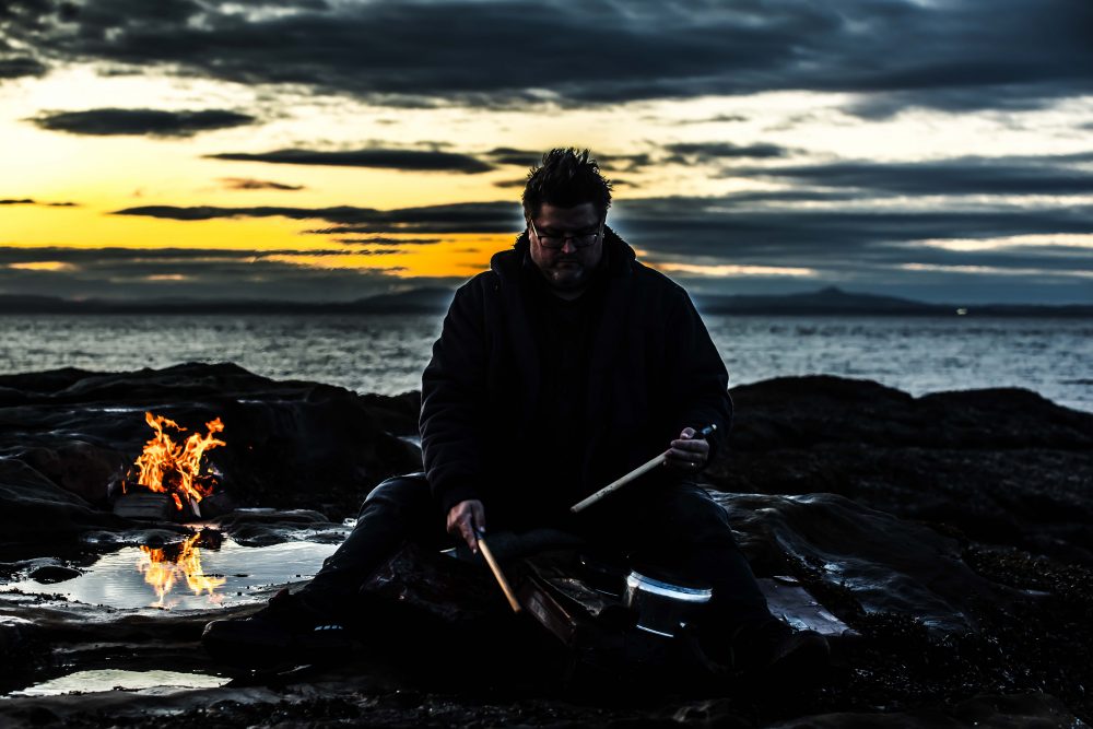 A man drumming on the beach next to a fire at dusk.