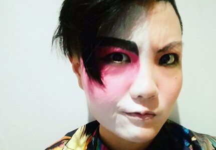 A photo of a person with an asymmetrical short haircut with theatrical make up on one half of their face. They are wearing a black and brightly coloured patterned shirt. 